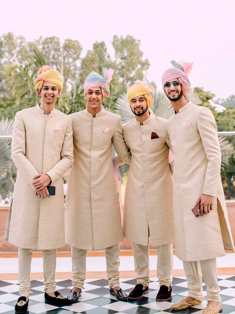 Four men in traditional cream-colored sherwanis and pastel turbans smiling together outdoors.