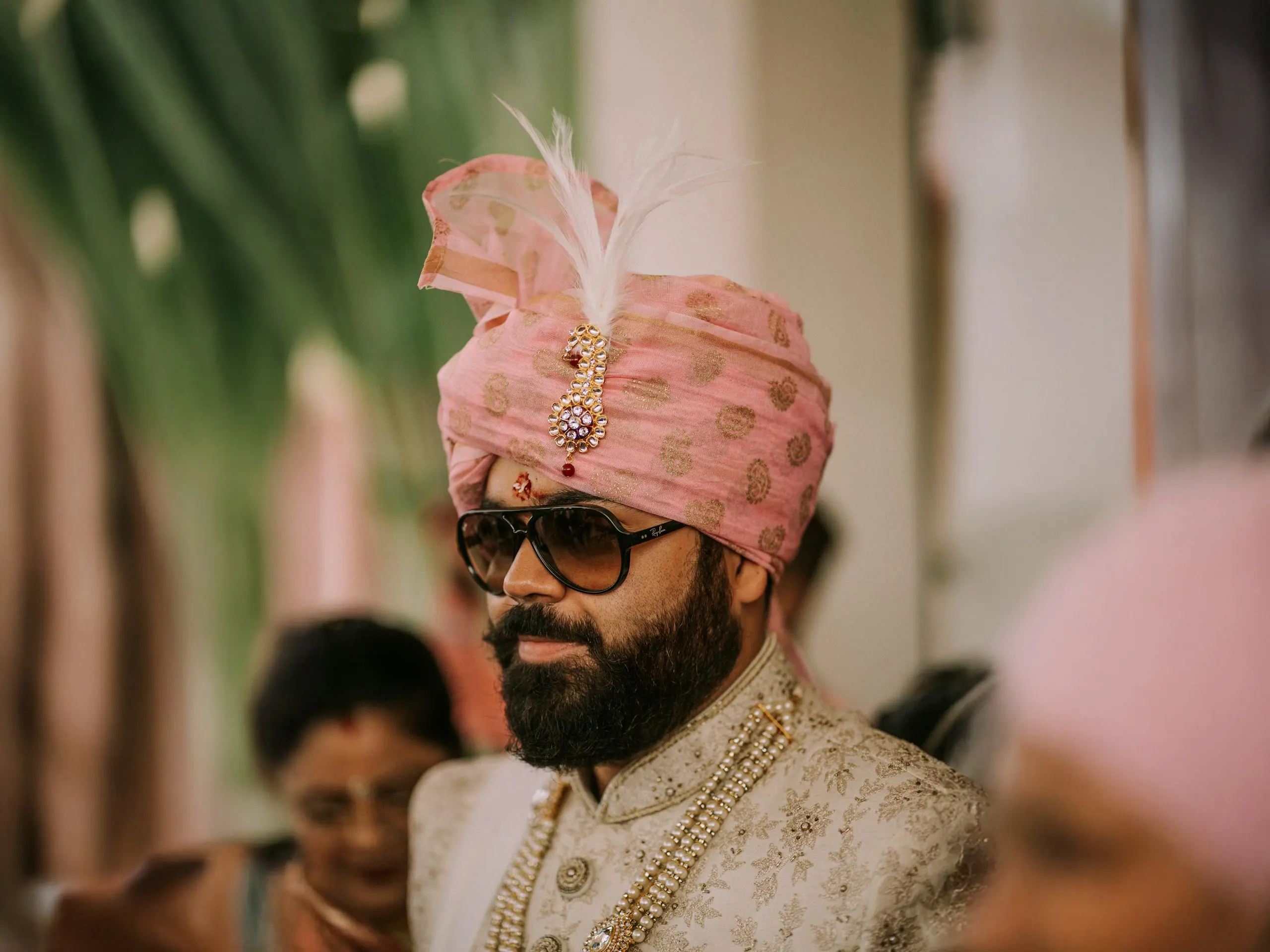 A man wearing a pink turban with a feather, sunglasses, and traditional attire at a wedding.